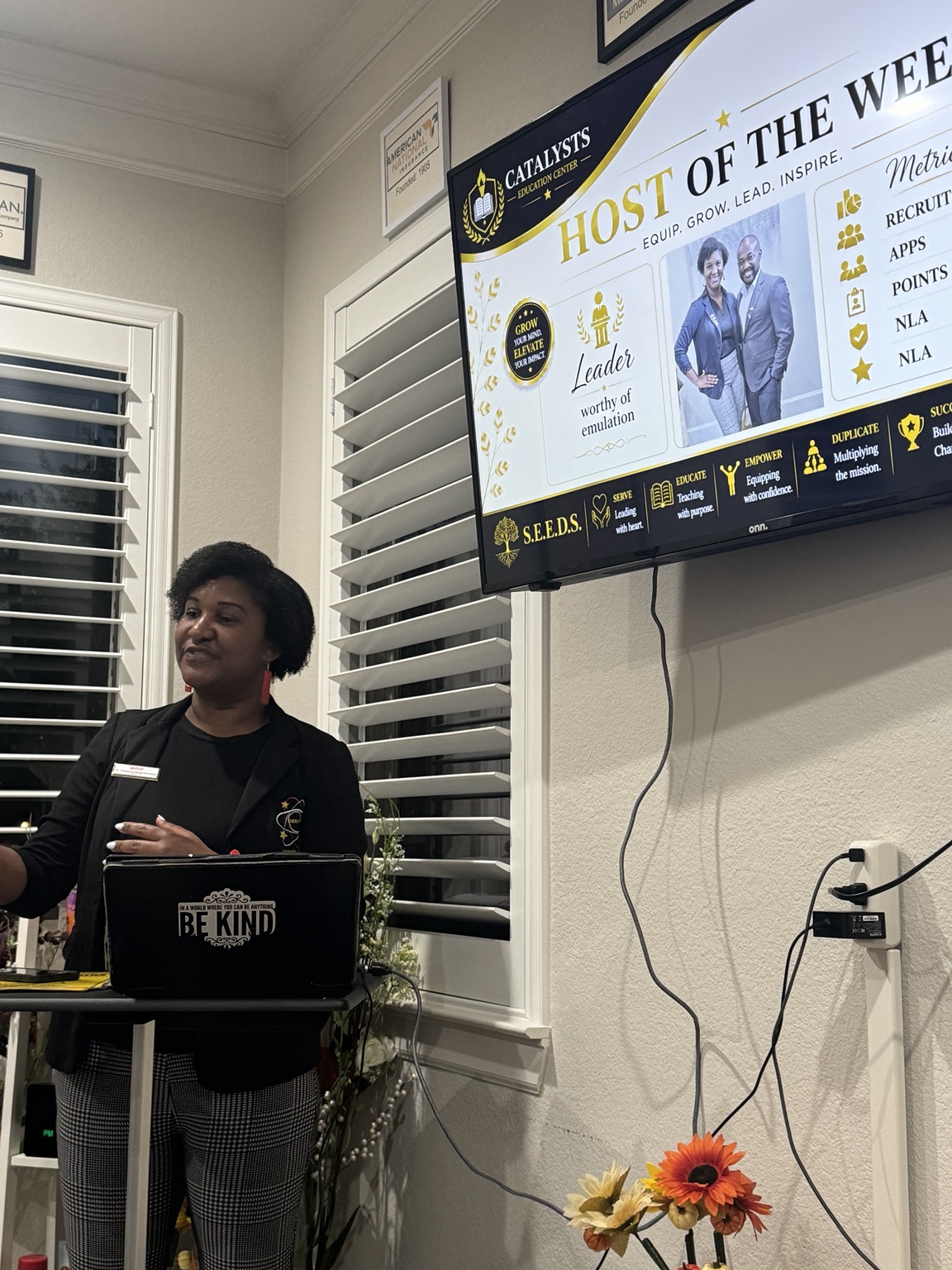 A presenter speaking at a lectern beside a wall screen showing a "Host of the Week" slide